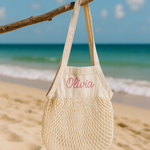 Beige mesh bag with personalised name embroidery on a beach with ocean in the background