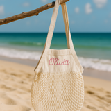 Beige mesh bag with personalised name embroidery on a beach with ocean in the background